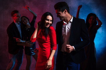 Hanging out with friends. Two young and attractive couples and one gorgeous woman are dancing together in the smoke and lights of the club, celebrating New Year’s Eve or Christmas.