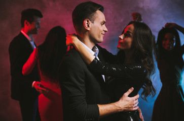 Falling in love. Gorgeous couple of smartly dressed man and a woman in black glittery dress is dancing in the dark, looking into each other’s eyes and smiling.