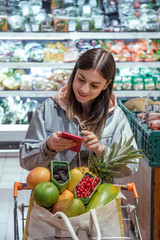 A young woman buys groceries in a supermarket with a phone in her hands.
