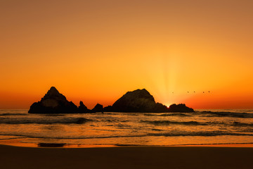Fototapeta na wymiar Sun rising over rock formations as pelicans fly over, waves