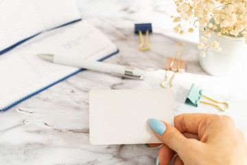 Woman's hand holds an empty business card in the office, in the background is a blue diary for scheduling meetings, a white pen and multicolored paper clips and white flowers on marble table.