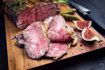 Traditional lunch meat with sliced cold cuts roast beef with gherkin and fig fruit as closeup on a modern design cutting board