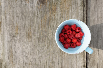 Top view of beautiful arrangement - ripe raspberries in blue mug, on old wooden board background. Copy space.