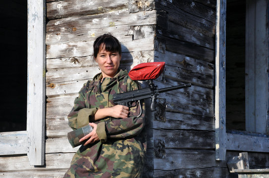 young woman in camouflage and paintball with a paintball marker near an abandoned building.