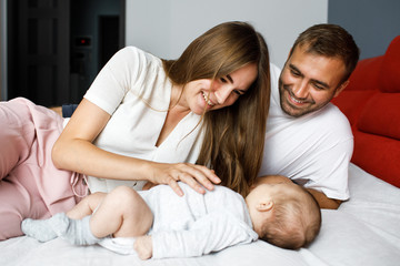 Young happy cheerful family of three lying in bed together. Parents play with their little baby