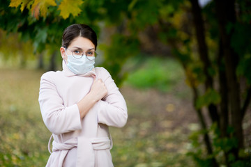 Young woman in a medical mask wrapped herself in a light coat on a cool autumn day