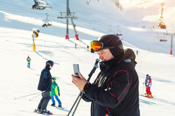 Senior mature skier making selfie with modern camera phone at winter alpine skiing resort. Old aged sporty person using smartphone at mountain Alps snow slope outdoors during cold sunny day