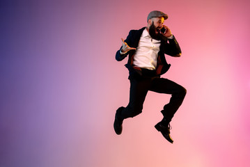 Obraz premium Full length portrait of happy jumping man wearing office clothes in neon light isolated on gradient background. Emotions, ad concept. Using smartphone, winning bet or sale, shopping, talking