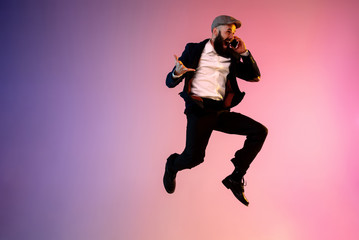 Fototapeta na wymiar Full length portrait of happy jumping man wearing office clothes in neon light isolated on gradient background. Emotions, ad. Using smartphone, winning bet or sale, shopping, hurrying up, office work.