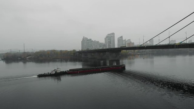 Small tugboat with scow is sailing on wide calm river in industrial smog. Major barge floating in fog on creek through concrete bridge at downtown