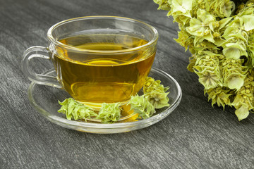 Greek traditional herbal tea in transparent cup. Bunch of mountain tea Malotira and mug with hot tea. Mountain tea sideritis. Natural Greek herbs on dark wooden background.