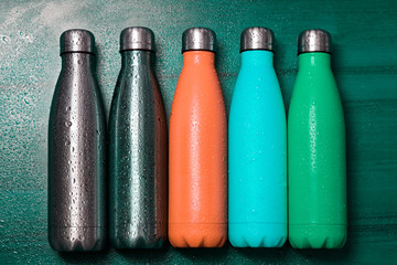Orange, cyan, green and silver eco steel thermo bottle for liquid on wooden table, sprayed with water.