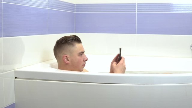 A young man lies in the bath and takes a selfie on the phone. A man relaxes in the bath.