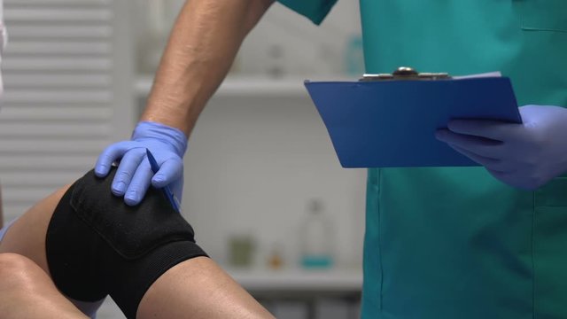 Traumatologist checking patients leg in knee brace, writing examination result