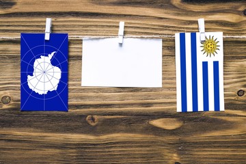 Hanging flags of Antarctica and Uruguay attached to rope with clothes pins with copy space on white note paper on wooden background.Diplomatic relations between countries.