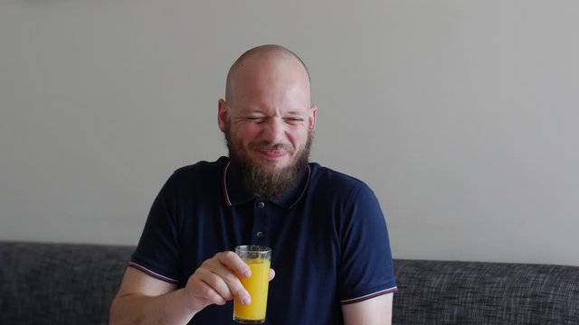 Handsome Man with beard drinking fresh Orange juice and disgusted by pulp. Funny face, navy blue shirt, sitting on apartment sofa.