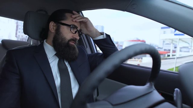 Bearded man suffering migraine sitting in car, stressful day, traffic jam