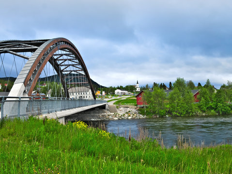 Bowstring Tied-Arch Bridge over the Glomma River at Alvdal, Norway on a cloudy evening with red barn and village buildings across the river.