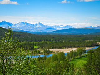 Naklejka na ściany i meble Beautiful farm valley landscape with a flowing river, snow capped mountains and green trees under a blue sky with fluffy clouds in Norway on a sunny summer day.