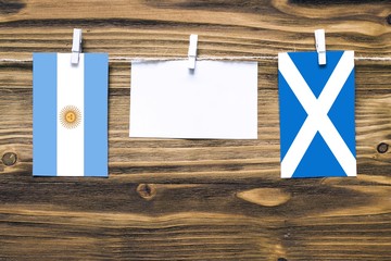 Hanging flags of Argentina and Scotland attached to rope with clothes pins with copy space on white note paper on wooden background.Diplomatic relations between countries.