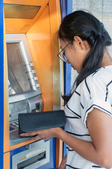Fototapeta na wymiar Young woman with wallet standing by ATM. Asian girl is withdrawing money from ATM on the street.