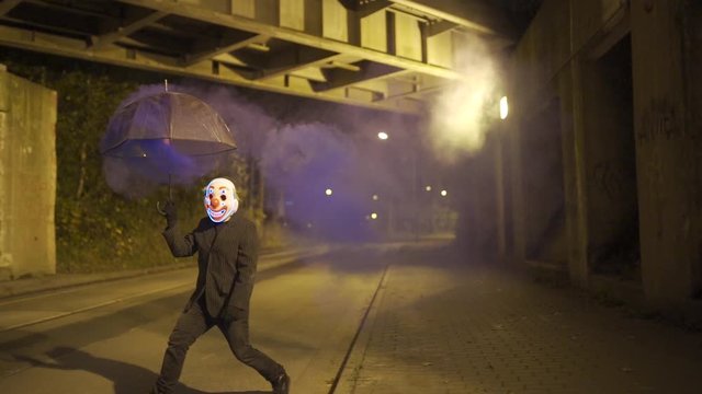 Man with clown mask and smoking umbrella is dancing under a bridge at night.
