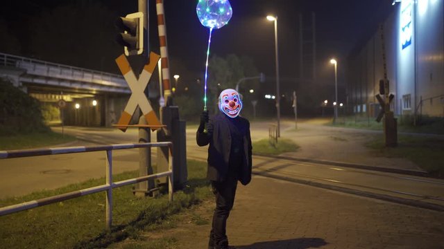 Clown with Joker Mask and glowing balloon is sitting near an railroad crossing.