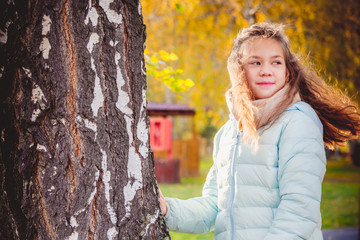 Smiling girl with disheveled hair in the wind in scarf and blue down jacket stands holding on to birch on an autumn day.