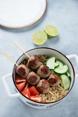Food bowl with meatballs on skewers, bulgur, quinoa and vegetables, elevated view over light-blue stone surface, vertical shot
