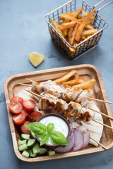 Fototapeta na wymiar Greek chicken souvlaki skewers with french fries, tzatziki and vegetables, vertical shot over grey concrete background