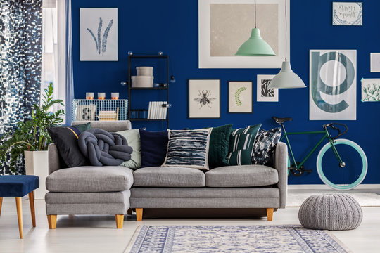 Grey and mint lamps above pouf and corner sofa with pillows in blue interior with posters on the wall