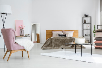 Classy white bedroom with king size bed, industrial furniture and trendy pink armchair, copy space on empty wall