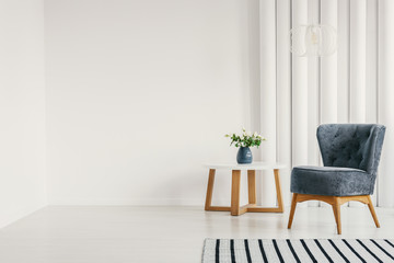 Fashionable blue armchair next to white round coffee table with vase with flowers in empty white interior with striped rug, real photo with copy space on empty white wall
