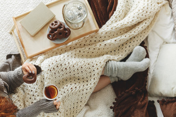 Top view of young girl covered with cozy white blanket siting on bed drinking tea and eating cookies