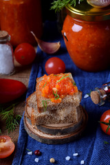 Adjika on a slice of rye bread and in a glass jar surrounded by the ingredients on the edge of the wooden table