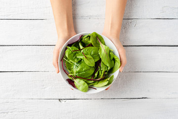 Overhead shot of woman’s hands holding green salad in bowl on white wooden background. Close up