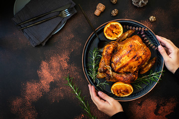 Baked whole chicken with oranges and rosemary. Female hands holding a tray with a festive Christmas dish on a dark rustic background. Top view, flat lay. Copyspace
