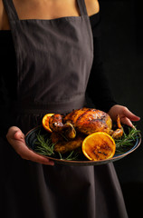 Baked whole chicken with oranges and rosemary. Girl in a rustic apron holding a tray with a festive Christmas dish. Dark background, low key. Vertical