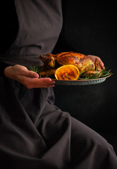 Baked whole chicken with oranges and rosemary. Girl in a rustic apron holding a tray with a festive...