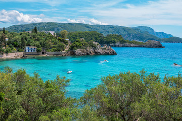 Landscape with turquoise calm sea water, mountain with rocky hillside covered with green trees and bushes and caves, cruise touristic boats and clouds on the sky. Corfu Island, Greece. 