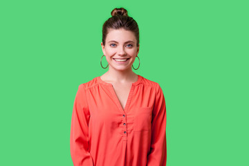 Portrait of lovely positive young woman with bun hairstyle, big earrings and in red blouse standing, looking at camera with toothy charming smile. indoor studio shot isolated on green background