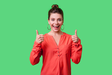 Thumbs up! Portrait of satisfied positive young woman with bun hairstyle, big earrings and in red blouse standing smiling widely, showing like gesture. indoor studio shot isolated on green background