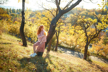 Fototapeta na wymiar Portraits of a charming red-haired girl with a cute face. Girl posing in autumn park in a sweater and a coral-colored skirt. In the hands of a girl a yellow leaf