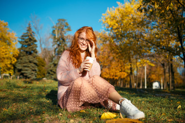 Portraits of a charming red-haired girl with glasses and a pretty face. Girl posing in autumn park in a sweater and a skirt of coral color.