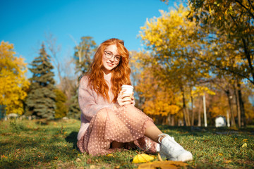 Portraits of a charming red-haired girl with glasses and a pretty face. Girl posing in autumn park in a sweater and a skirt of coral color. - obrazy, fototapety, plakaty