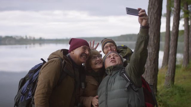 Happy senior hikers smiling, showing bunny ears and posing together for smartphone camera while taking selfie against beautiful lake on autumn day