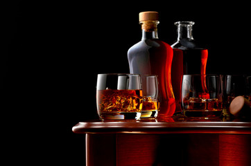 Two glasses of whiskey on a wooden table. In the background, two bottles of whiskey and two glasses of whiskey. Low key.