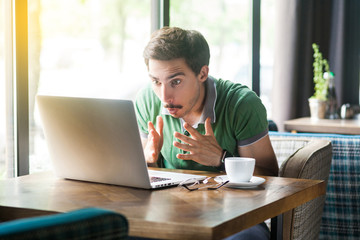 Fototapeta na wymiar Oh my God! Young shocked businessman in green t-shirt sitting, looking at some unbelievable news at laptop screen and surprised. business and freelancing concept. indoor shot near window at daytime.