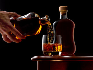 A glass of whiskey on the rocks and a full bottle of whiskey on the wooden table. The glass is...