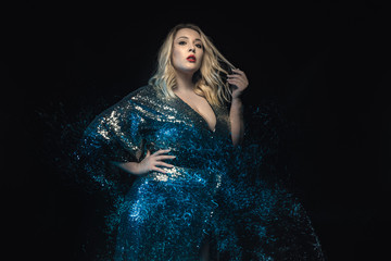 Half length plus size model shot with mixed lights effect. Black background. Young blonde woman in shiny evening dress posing towards the camera. Fashion glamour shooting.